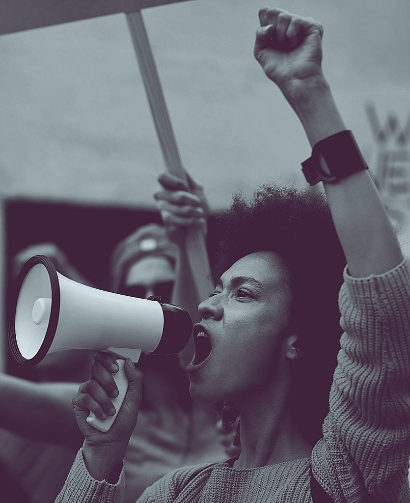 Black and white photo of a person at a protest holding a megaphone with their fist held high.