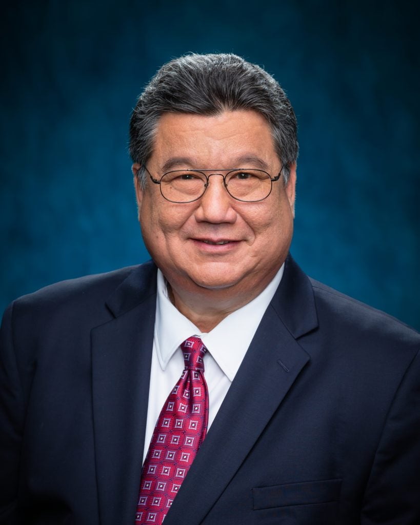 Headshot of Ron Kouchi smiling in front of a blue background.